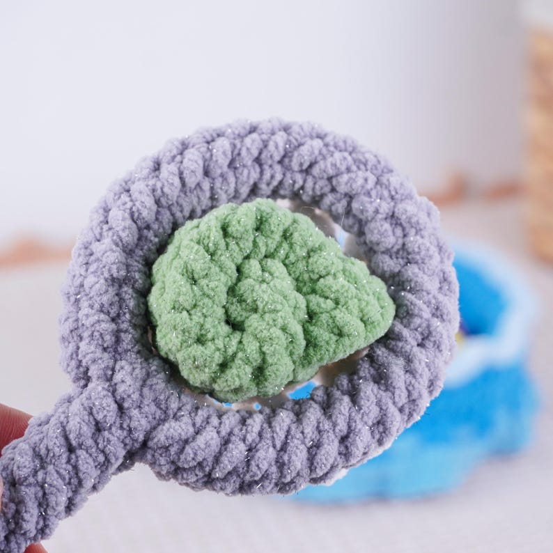 May include: A handmade crochet toy in a whimsical design. The toy features a light purple ring with a green center, creating a soft, textured appearance. The plush material suggests a focus on comfort and tactile appeal, ideal for children's play.