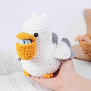 May include: A handmade white crochet pelican with gray wings, a yellow pouch, and orange feet. The pelican has a black eye and is holding a small blue object in its pouch. This soft toy is a unique, handcrafted item.