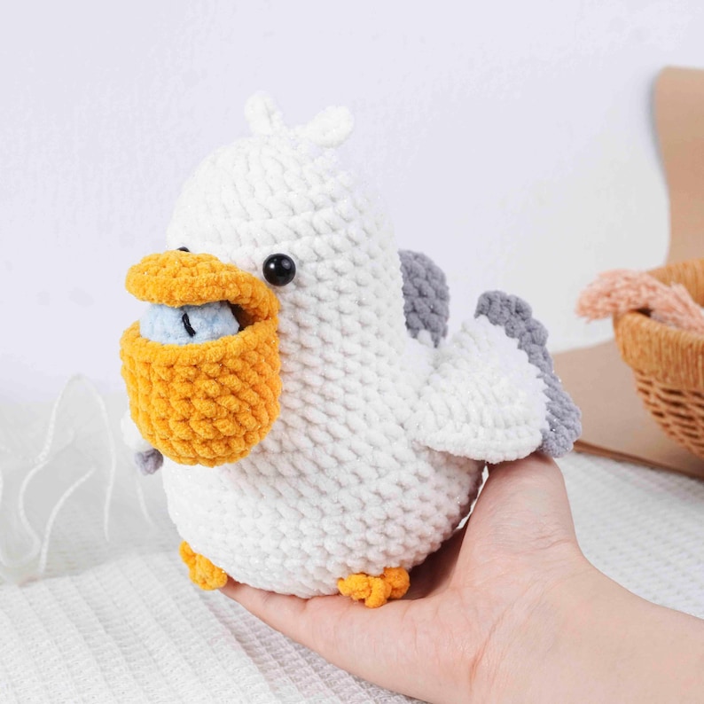 May include: A handmade white crochet pelican with gray wings, a yellow pouch, and orange feet. The pelican has a black eye and is holding a small blue object in its pouch. This soft toy is a unique, handcrafted item.