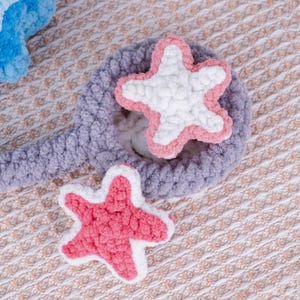 May include: Crocheted starfish in white, pink, and lavender. The starfish are placed on a lavender crocheted circle with a handle. The background is a textured, light brown fabric.
