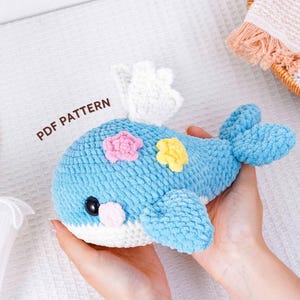 May include: A handmade blue crochet whale with white, pink, and yellow accents. The whale has a white belly, black eyes, and a white fin. The text "PDF PATTERN" is visible. This soft toy is perfect for a nursery or as a decorative item.