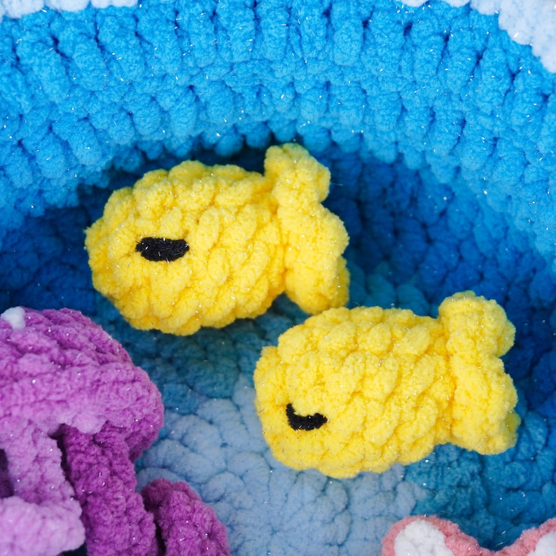 May include: Close-up of a handmade crochet scene featuring two yellow fish with black eyes. The fish are set against a backdrop of blue and white crochet, with a purple jellyfish and pink object in the foreground. The scene evokes an underwater theme.