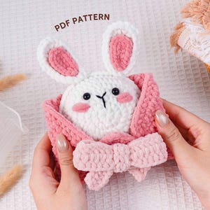 Può includere: Un giocattolo a forma di coniglietto all'uncinetto fatto a mano con corpo bianco, dettagli rosa e un fiocco. Il coniglietto ha occhi neri e guance rosa. Le orecchie sono bianche con l'interno rosa. Il giocattolo è avvolto in una coperta rosa. Il testo "PDF PATTERN" è visibile.