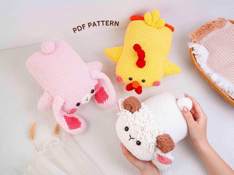 Op de afbeelding: Drie gebreide knuffels: een roze konijn, een gele kuiken en een wit lam. Het konijn heeft roze oren, de kuiken een rode kam en het lam bruine oren. De tekst "PDF PATTERN" is zichtbaar.