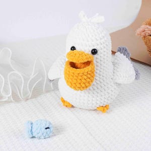 May include: A handmade white crochet pelican with a yellow pouch, gray wings, and orange feet. A small blue crochet fish is next to the pelican. The pelican has black eyes.