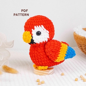 Può includere: Un pappagallo fatto a mano all'uncinetto, nei colori rosso, giallo, bianco e blu. L'uccello ha una testa rotonda, un becco giallo e un occhio nero. Il testo "PDF PATTERN" è visibile. Il pappagallo è montato su una piccola base di legno.