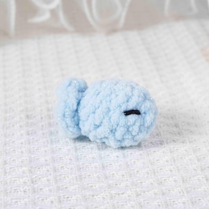 May include: A small, light blue crocheted fish with a round body and a small tail fin. The fish has a black stitched eye and is made of a soft, textured yarn with subtle glitter accents. The handmade item is likely a toy or decorative item.