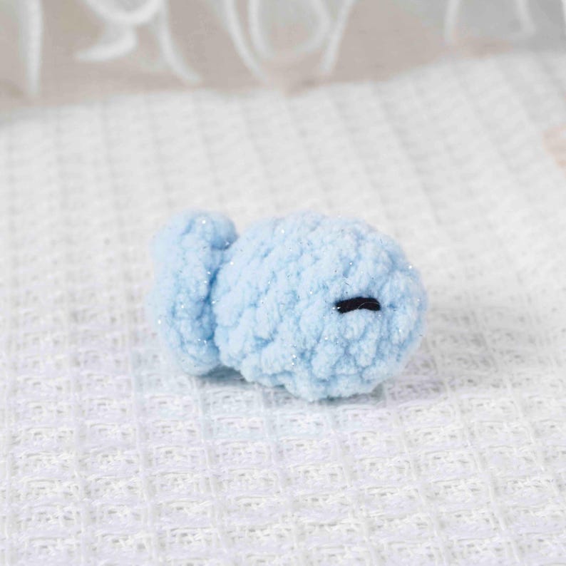 May include: A small, light blue crocheted fish with a round body and a small tail fin. The fish has a black stitched eye and is made of a soft, textured yarn with subtle glitter accents. The handmade item is likely a toy or decorative item.