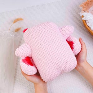Op de afbeelding: Een handgemaakt roze gebreid pluche speelgoed met rode accenten op de oren en voeten. Het zachte speelgoed wordt in twee handen gehouden en toont zijn textuur en knuffelige ontwerp. Het speelgoed is lichtroze van kleur.