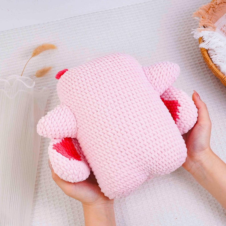 Op de afbeelding: Een handgemaakt roze gebreid pluche speelgoed met rode accenten op de oren en voeten. Het zachte speelgoed wordt in twee handen gehouden en toont zijn textuur en knuffelige ontwerp. Het speelgoed is lichtroze van kleur.