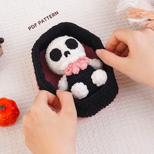 Pode incluir: Uma boneca de crochê com cabeça de caveira branca, órbitas oculares pretas e gola de babados rosa, descansando dentro de um caixão de crochê preto. O texto PDF PATTERN é visível acima do caixão. A boneca tem corpo preto com mãos e pés brancos.