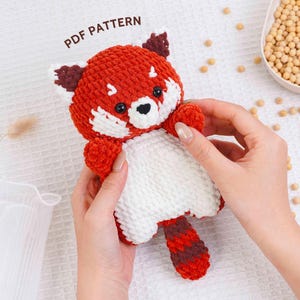 May include: A crocheted red panda toy with white details and a striped tail. The plushie is held by hands and has the text "PDF PATTERN" above it. The red panda has black eyes and a black nose.