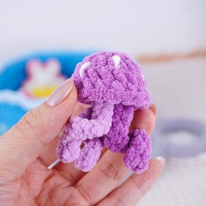 May include: A handmade, lavender-colored crochet jellyfish with white accents. The plush toy features a rounded head and flowing tentacles, showcasing a soft, textured design. This whimsical creation is a charming addition to any collection.