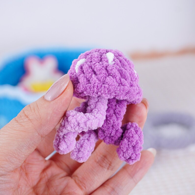 May include: A handmade, lavender-colored crochet jellyfish with white accents. The plush toy features a rounded head and flowing tentacles, showcasing a soft, textured design. This whimsical creation is a charming addition to any collection.
