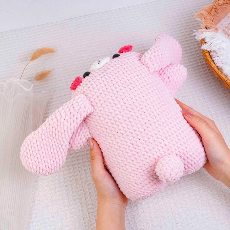 Op de afbeelding: Een handgemaakt roze pluche speelgoed, dat lijkt op een stripfiguur, met grote oren, een kleine staart en zwarte en rode accenten. Het speelgoed is gemaakt van een zacht, gebreid materiaal.