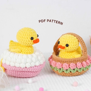 May include: Two handmade crochet ducklings, one in a pink, white, and pink egg-shaped container, and the other in a brown basket with pink and green floral trim. The ducklings are yellow with orange beaks and black eyes. The text "PDF PATTERN" is above the objects.