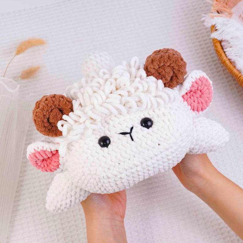Op de afbeelding: Een handgemaakt, wit gehaakt schaap knuffel met bruine oren en accenten. Het schaap heeft zwarte ogen en een eenvoudig gestikt gezicht. De oren hebben roze binnendetails. De knuffel is zacht en knuffelig, perfect als kinderspeelgoed of voor de kinderkamerdecoratie.