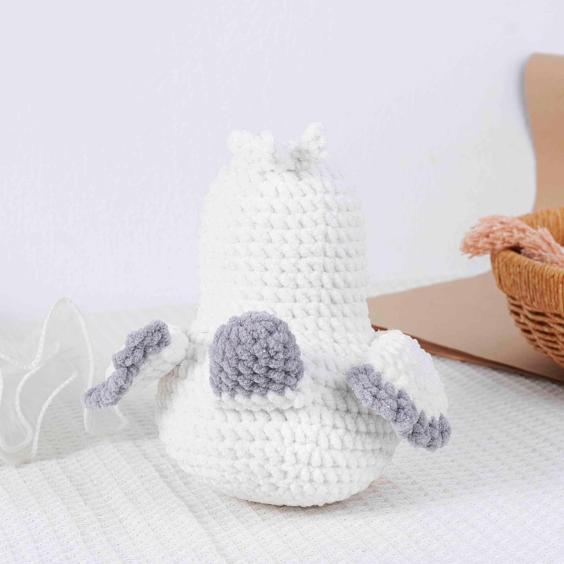 May include: A handmade white crochet bird with gray accents. The bird has a rounded body, small wings, and a gray pom-pom on its back. The bird is sitting on a white surface.