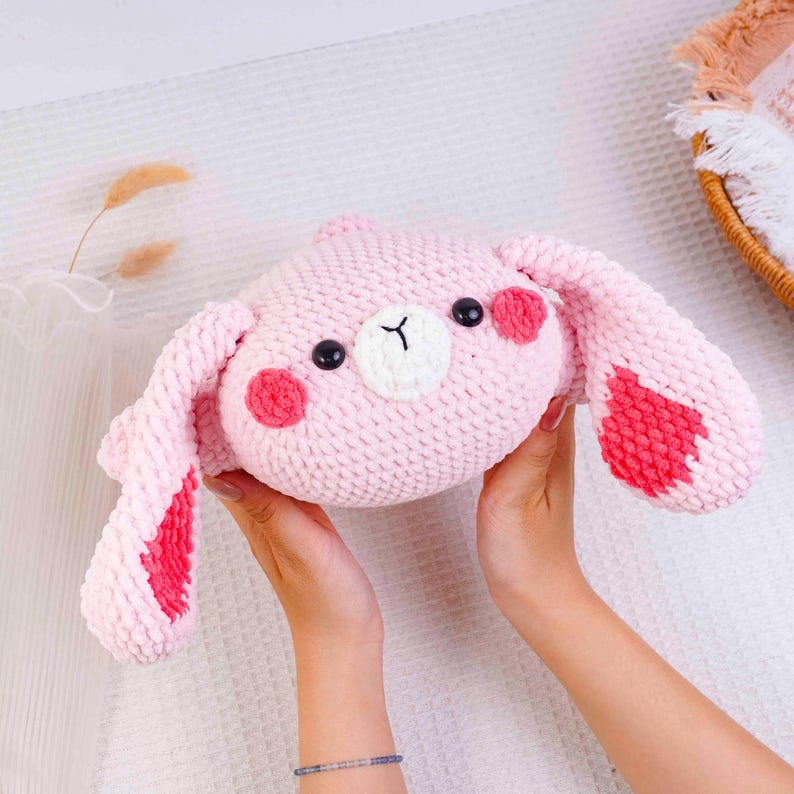 Op de afbeelding: Een handgemaakte roze gehaakte konijn knuffel met zwarte knoopogen, een witte neus en rode accenten op de wangen en oren. Het konijn wordt door twee handen omhoog gehouden. De knuffel is een zacht, knuffelig speelgoed.