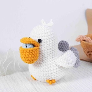 May include: A handmade white crochet bird with a yellow beak holding a blue ball. The bird has gray wings and orange feet. The bird is sitting on a white surface.