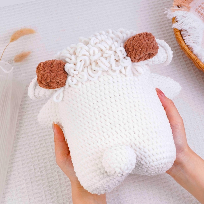 Op de afbeelding: Een handgemaakt wit gehaakt schaap speelgoed met bruine oren en een pluizige witte wollige kop. Het speelgoed wordt in de handen van een persoon gehouden. Het schaap heeft een kleine staart en is gemaakt van zacht garen.