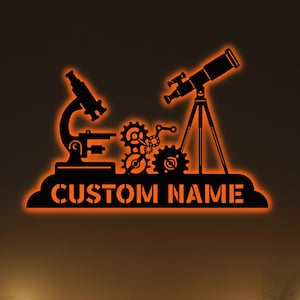 Puede incluir: Arte de pared de metal negro con un microscopio, un telescopio y engranajes, iluminado con un brillo naranja. Las palabras "CUSTOM NAME" están debajo. Regalos y luces de cadena en primer plano, creando un ambiente festivo.