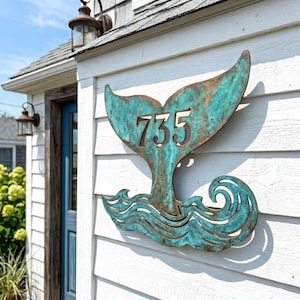 May include: A metal house number sign shaped like a whale tail with stylized waves at the bottom. The sign features a weathered teal and copper patina finish and displays the numbers 735 in a dark, cut-out font centered on the whale tail.