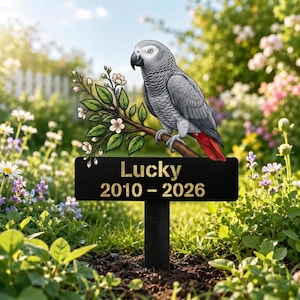 May include: A black metal garden stake featuring a grey African Grey parrot with a red tail perched on a flowering branch. Below the bird, a rectangular plaque displays the text Lucky and the years 2010 - 2026 in a gold-coloured font.