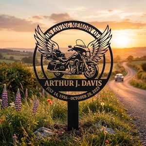 Könnte beinhalten: Ein rundes Gedenkschild aus schwarzem Metall mit einem Motorrad mit großen Flügeln, dem Text IN LOVING MEMORY OF, dem Namen ARTHUR J. DAVIS und den Daten JULY 14, 1956 - OCTOBER 28, 2023, montiert auf einem vertikalen Stab zur Bodenbefestigung.