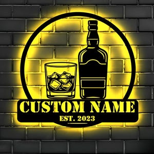 May include: Black metal sign with a whiskey bottle and a glass of ice cubes inside a circle, with the words "CUSTOM NAME" and "EST. 2023" below. The sign is illuminated with a yellow glow against a brick wall.
