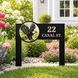 Può includere: Segnaletica stradale in metallo nero con un'aquila intagliata. Il cartello mostra il numero "22" e "CANAL ST." in lettere dorate. Il cartello è posto in un giardino con fiori e vegetazione.