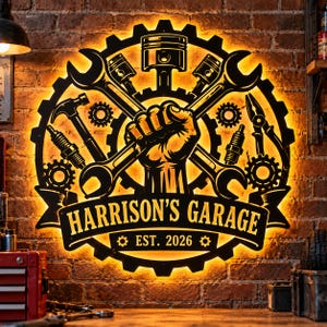 May include: A circular black metal wall sign with warm backlighting featuring a central fist holding a wrench, surrounded by gears, a piston, a hammer, spark plugs, and pliers. The banner reads HARRISON'S GARAGE and EST. 2026 in bold text.