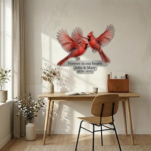 Puede incluir: Decoración de pared con dos pájaros cardenales rojos con las alas extendidas. Las aves están colocadas cerca una de la otra, con el texto "Forever in our hearts John & Mary 1970-2023" debajo. La obra de arte está montada en una pared clara.