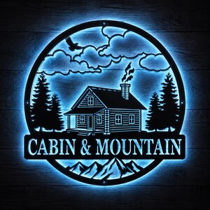 May include: A black metal sign with a blue LED backlight. The sign features a cabin, mountains, trees, and an eagle silhouette within a circular frame. The words "CABIN & MOUNTAIN" are displayed at the bottom.