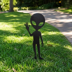 May include: A black metal silhouette of an alien figure with a large oval head, almond-shaped eyes, and a slender body. The figure is depicted in a standing position with one arm raised in a waving gesture, featuring a flat, cutout design.