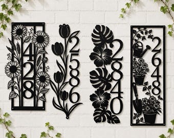 May include: Black metal address signs with floral designs. The signs feature numbers and decorative elements like sunflowers, tulips, hibiscus, and gardening tools. The signs are mounted on a white brick wall.