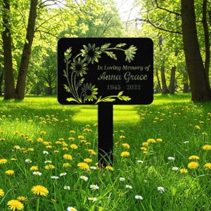 May include: A black metal memorial garden stake with a rectangular sign. The sign features floral designs and the inscription "In Loving Memory of Anna Grace 1945-2022" in green lettering. The stake is placed in a grassy area with wildflowers.