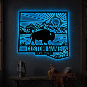 Pode incluir: Uma peça de arte de parede em metal preto com uma silhueta de bisonte, montanhas e um design de sol, iluminada por uma luz LED azul. O texto "CUSTOM NAME" e "EST. 2026" estão incluídos. A obra de arte é exibida em uma parede escura.