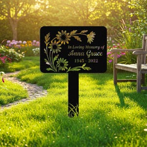 May include: Black metal memorial garden stake with gold floral design and the text "In Loving Memory of Anna Grace 1945-2022". The stake is rectangular with rounded corners and is placed in a grassy area.