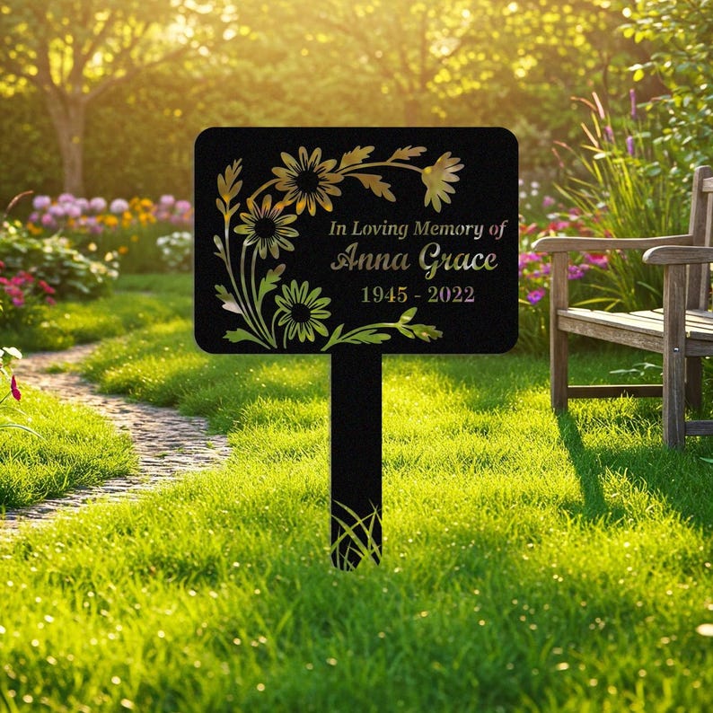 May include: Black metal memorial garden stake with gold floral design and the text "In Loving Memory of Anna Grace 1945-2022". The stake is rectangular with rounded corners and is placed in a grassy area.