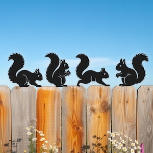 Op de afbeelding: Zwarte metalen silhouetten van eekhoorns op een houten hek tegen een blauwe lucht. De eekhoorns staan in verschillende poses, met pluizige staarten en gedetailleerde kenmerken. Madeliefjes en witte bloemen aan de voet van het hek.