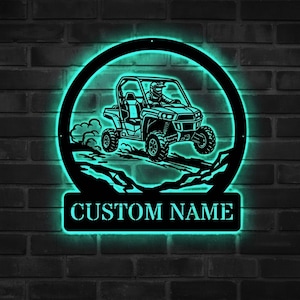 Könnte beinhalten: Schwarze Metall-Wandkunst mit einem Geländewagen mit Fahrer, eingebettet in einen kreisförmigen Rahmen mit türkisfarbenem Leuchten. Die Worte "CUSTOM NAME" sind darunter abgebildet. Das Kunstwerk ist an einer dunklen Backsteinmauer angebracht.