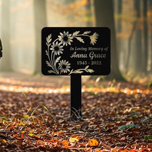 May include: Black metal memorial garden stake with floral accents. The sign reads "In Loving Memory of Anna Grace 1945-2022". The stake is rectangular with a decorative floral design and a black finish. The background is a blurred forest scene.