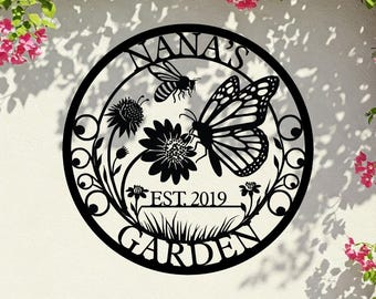 May include: Black metal garden sign with the text "NANA'S GARDEN EST. 2019". The sign features a bee, a butterfly, flowers, and grass within a circular frame. Decorative garden art.