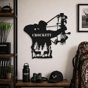 Peut inclure: Décoration murale en métal noir représentant un archer avec un arc et des flèches, avec le nom "CROCKETT". Le design comprend des silhouettes d'arbres et de cerfs. Une gourde noire, des jumelles et une casquette avec un logo de cerf sont sur une surface en bois.
