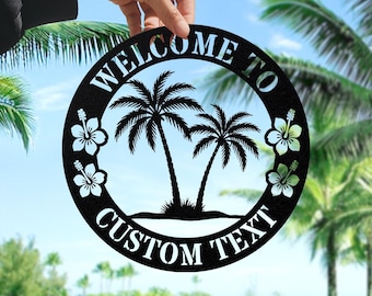 Könnte beinhalten: Schwarzes Metall-Willkommensschild mit kreisförmigem Design. Das Schild zeigt Palmen und Hibiskusblüten. Die Wörter "WELCOME TO" und "CUSTOM TEXT" sind aus dem Metall ausgeschnitten. Das Schild wird von einer Hand gehalten.