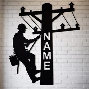 May include: Black metal wall art depicting a lineman working on a utility pole. The silhouette includes the word "NAME" and small house-shaped cutouts. The artwork is mounted on a white brick wall.