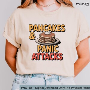 Può includere: Una maglietta beige con il testo "PANCAKES & PANIC ATTACKS" in arancione e rosso. Un'immagine di cartone animato di una pila di pancake con burro e sciroppo è sopra il testo.