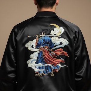 Puede incluir: Chaqueta bomber de satén negro con un diseño bordado en la espalda. El diseño presenta un guerrero samurái con atuendo azul y rojo, sosteniendo dos espadas, sobre un fondo de nubes y una luna creciente.