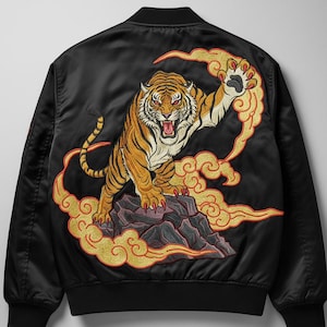 Puede incluir: Chaqueta bomber negra con un diseño de tigre bordado. El tigre naranja y negro está sobre una roca con nubes doradas y rojas. La chaqueta tiene cuello, puños y dobladillo de canalé.